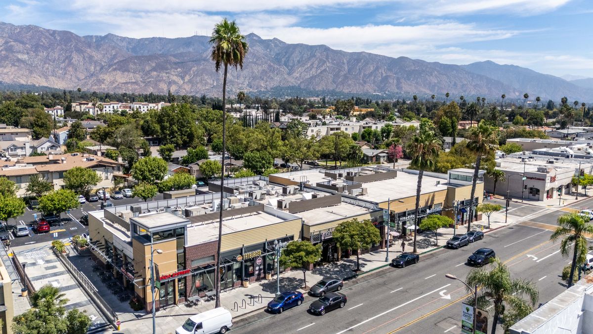Elevated view of the main building at Campus Village from the west, with cars on Colorado Boulevard, palm trees, and the mountains behind.