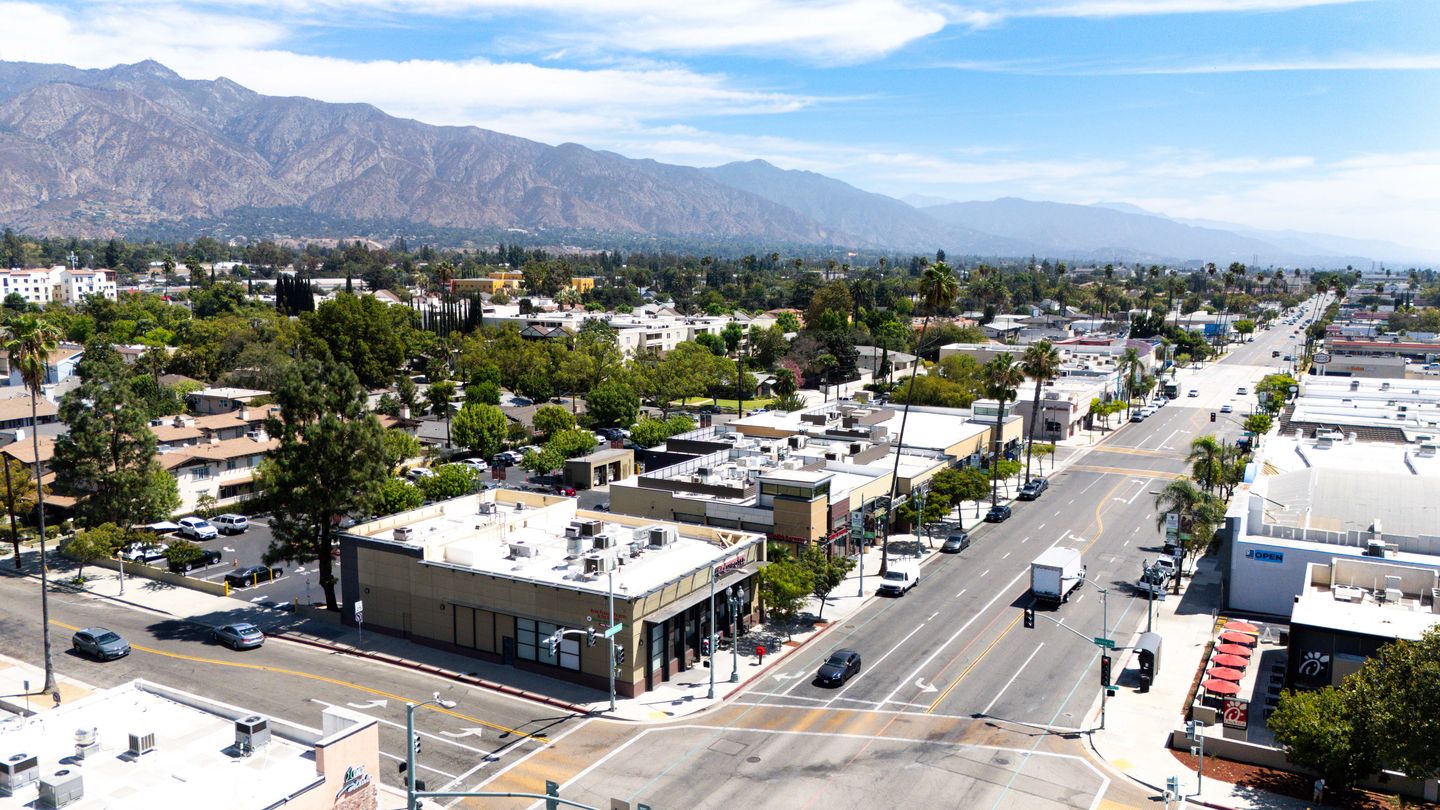 Aerial view looking east along Colorado Boulevard in Pasadena, with the Campus Village corner building at Bonnie Avenue in the foreground, the main building beyond it, and the San Gabriel Mountains behind.
