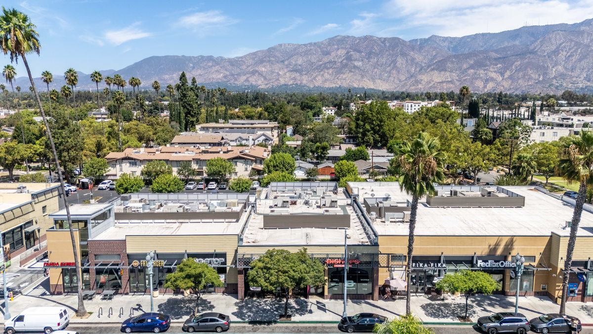 The main building at Campus Village from above Colorado Boulevard, with storefront signs along the row and the San Gabriel Mountains behind.