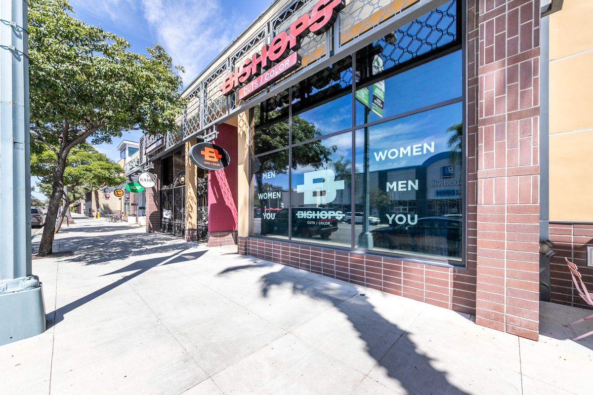 Suite 1 storefront on Colorado Boulevard, the former Bishops hair salon, with brick-clad columns, a metal canopy, and a full-height glass front under street trees.